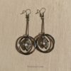 RAGNISH Ethereal Orbit Earrings. Gunmetal drop earrings with double hoop and suspended rhombus-cut clear crystal on beige fabric.