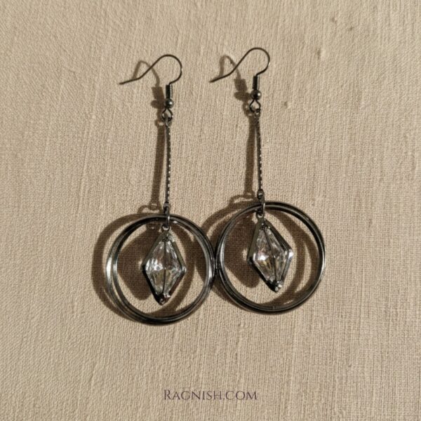 RAGNISH Ethereal Orbit Earrings. Gunmetal drop earrings with double hoop and suspended rhombus-cut clear crystal on beige fabric.