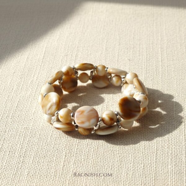 RAGNISH Amber Tide bracelet, two-strand design with polished, marbled caramel and cream oval beads on a textured beige surface.